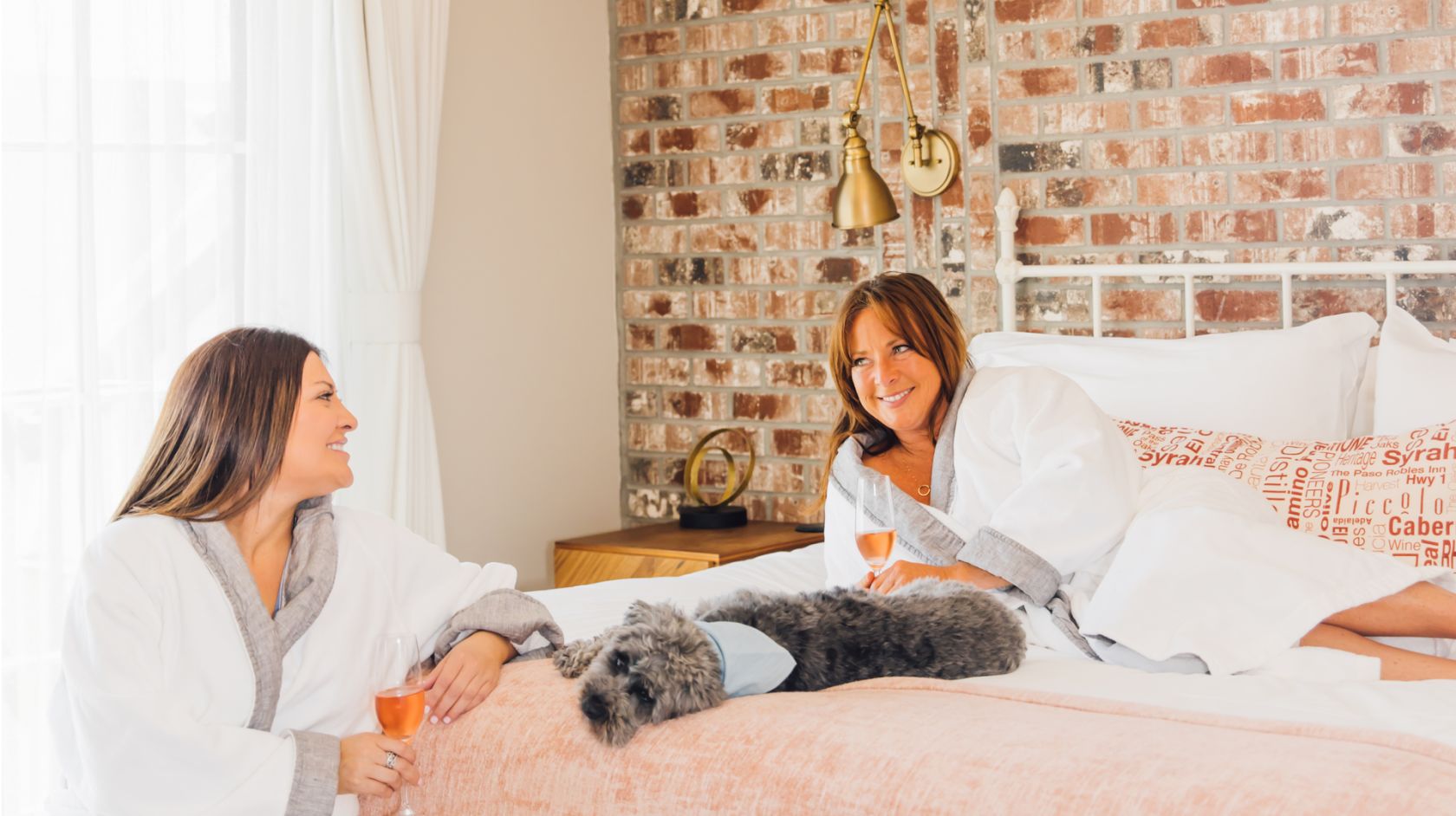 A Couple Of Women Sitting On A Bed With A Cat And A Glass Of Wine